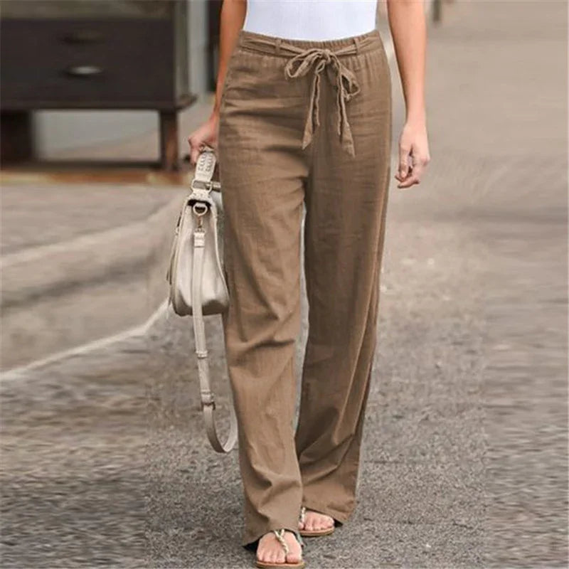 Woman wearing brown drawstring linen pants with white top, carrying beige handbag on city street