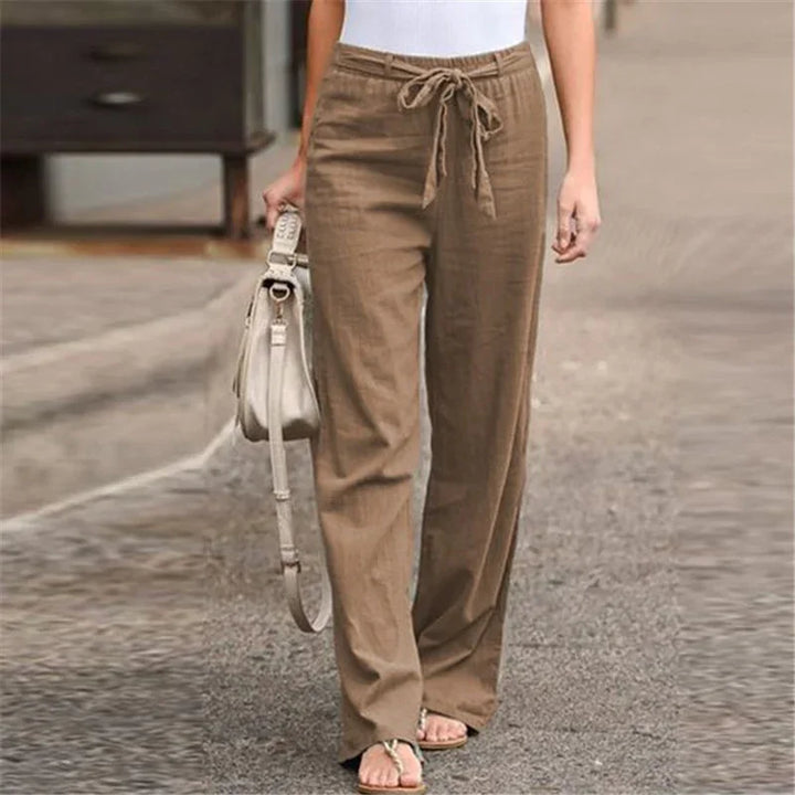Woman wearing brown drawstring linen pants with white top, carrying beige handbag on city street