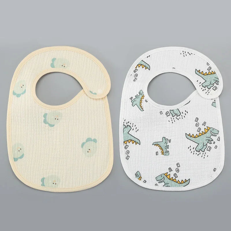 Two baby bibs on gray background, one beige with duck prints, one white with green dinosaur prints