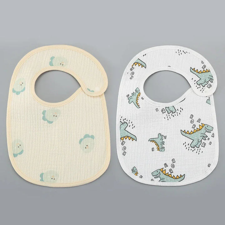 Two baby bibs on gray background, one beige with duck prints, one white with green dinosaur prints