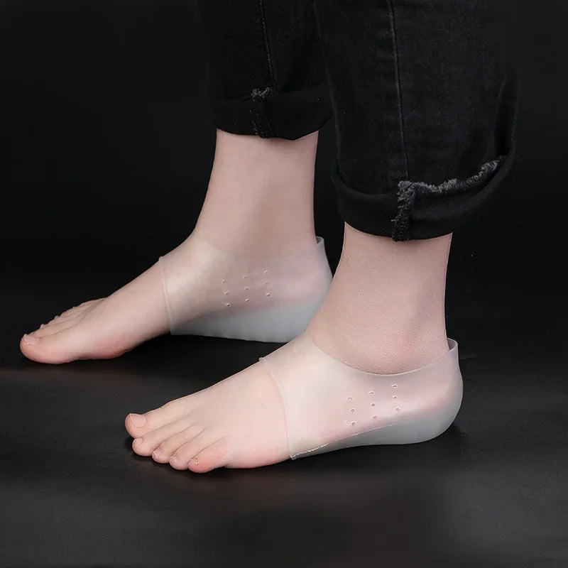Feet wearing transparent silicone heel protectors on black floor with cuffed black jeans