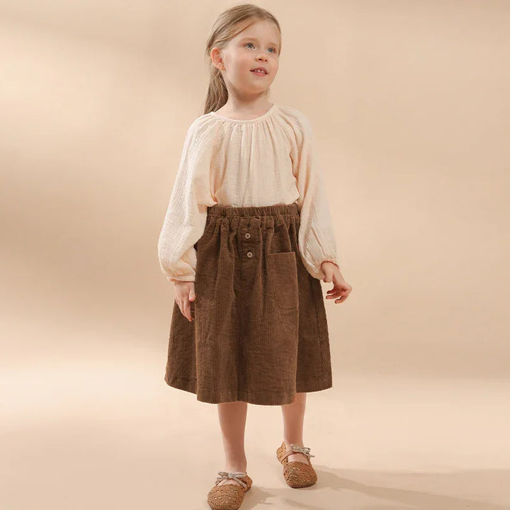 Young girl wearing a beige long-sleeve top and brown corduroy skirt with front pocket on beige background