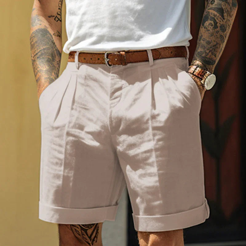 Man wearing beige pleated shorts with rolled cuffs, brown leather belt, white t-shirt, and tattooed arms