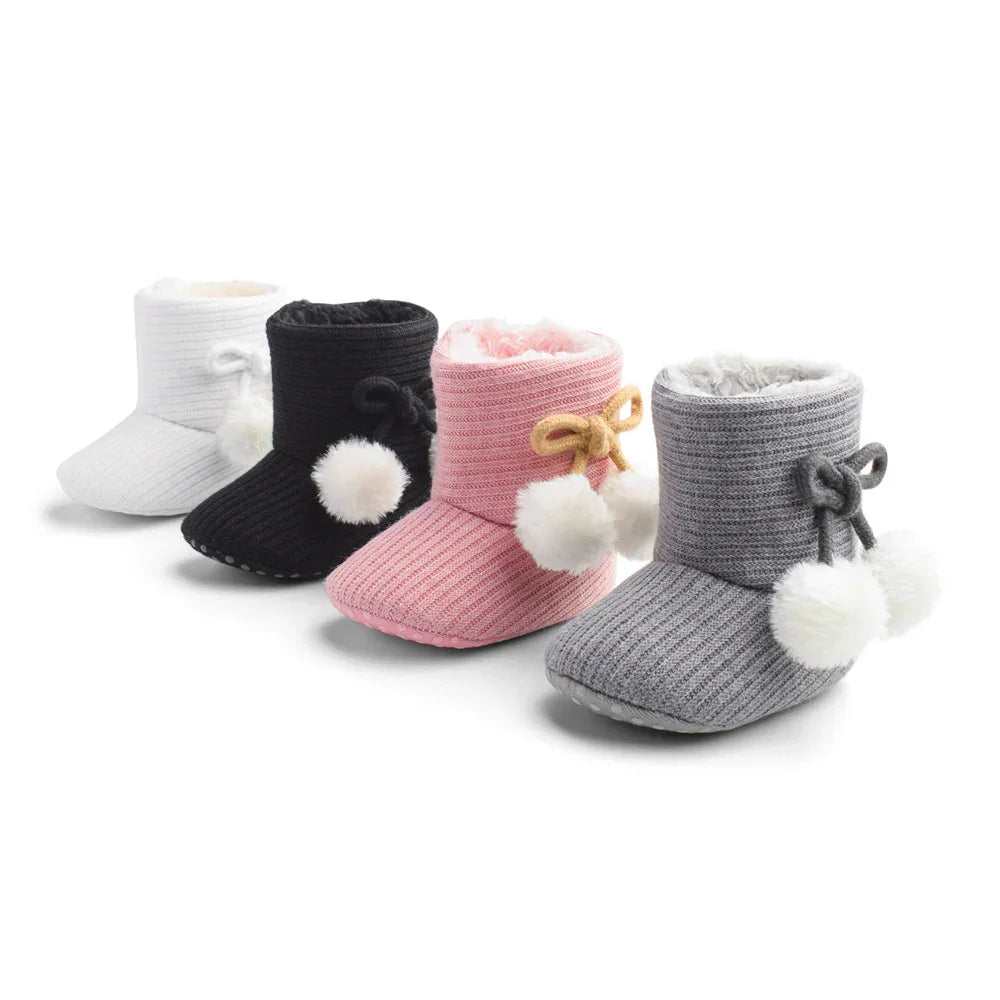 Four pairs of cozy knit baby booties in white, black, pink, and gray with pom-poms and bows
