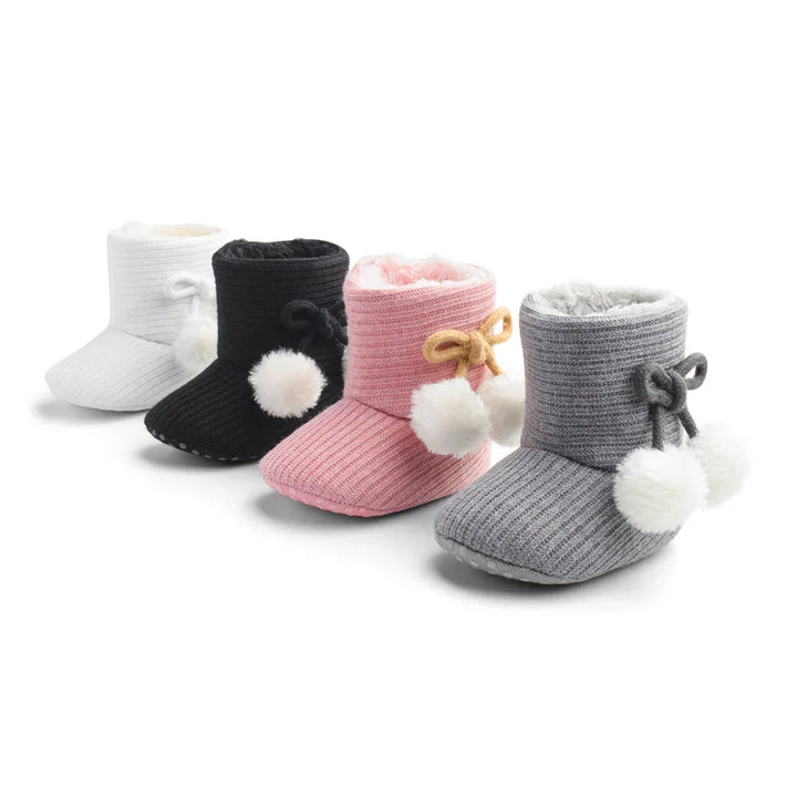 Four pairs of cozy knit baby booties in white, black, pink, and gray with pom-poms and bows