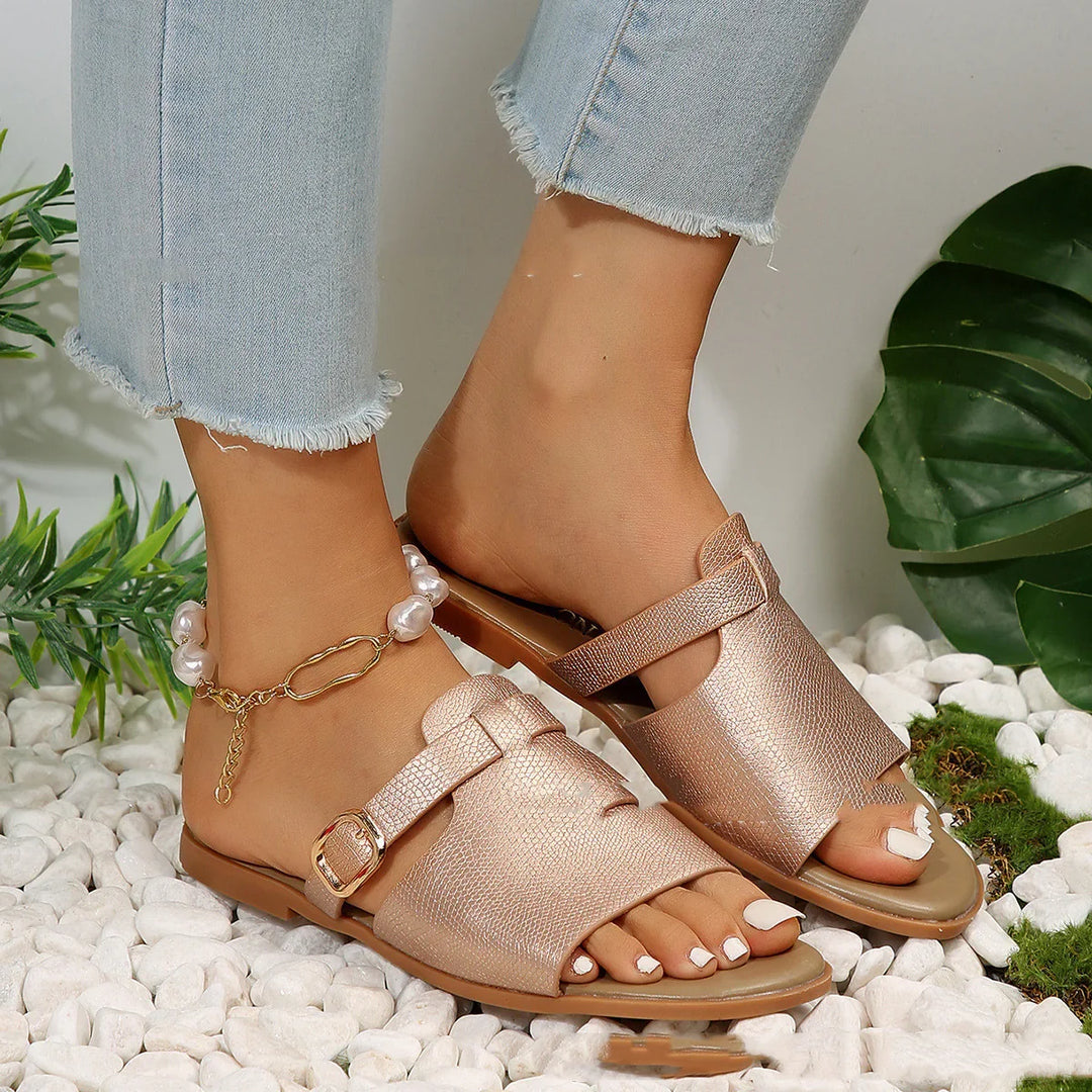 Close-up of feet wearing rose gold open-toe flat sandals with buckle, paired with cropped frayed jeans and pearl anklet