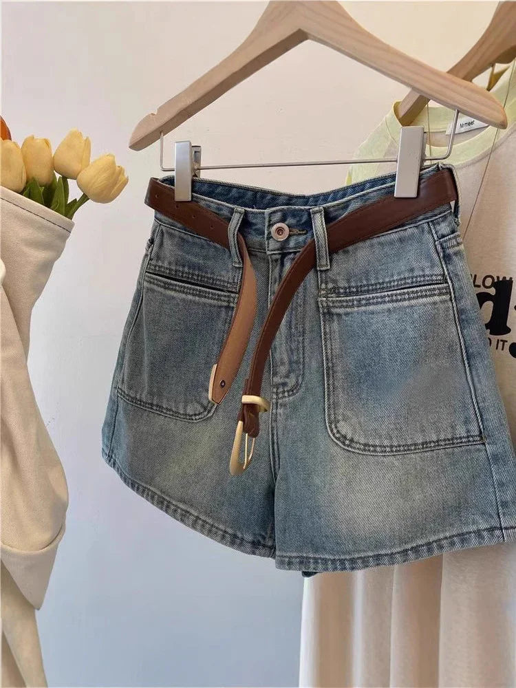 Denim shorts with front pockets and brown belt hanging on wooden hanger against white background