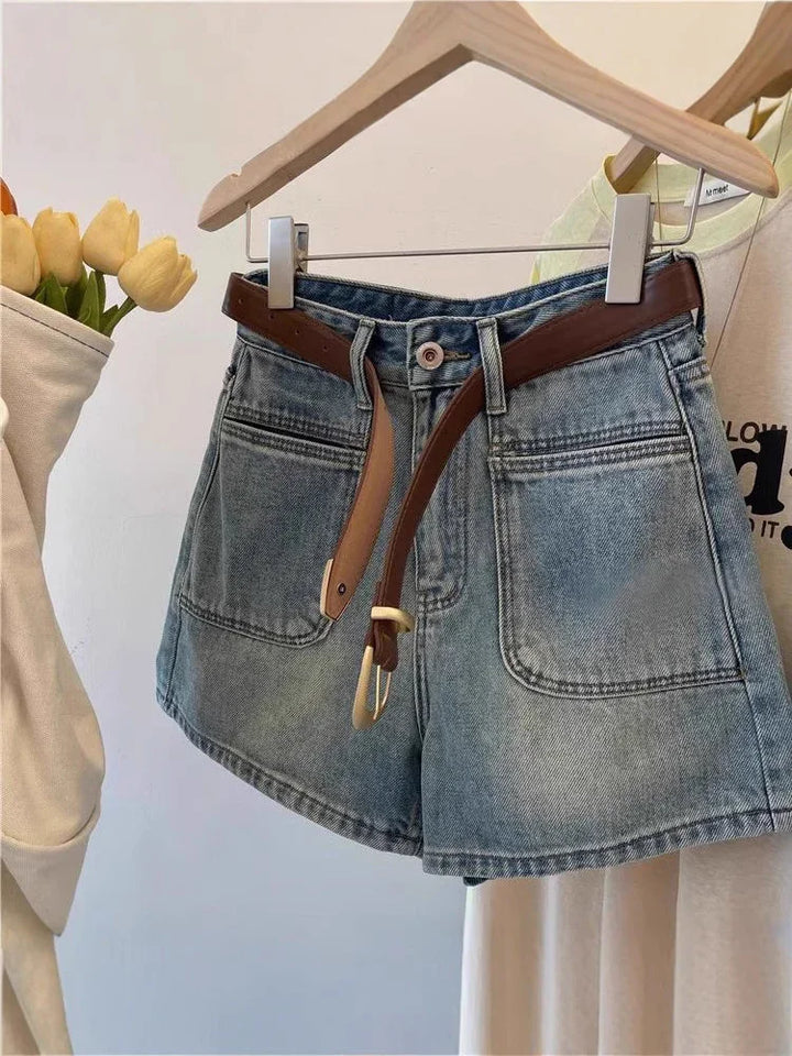 Denim shorts with front pockets and brown belt hanging on wooden hanger against white background