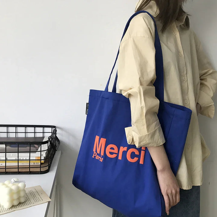 Person wearing beige shirt carrying blue Merci Paris tote bag indoors near desk with books and decorative items