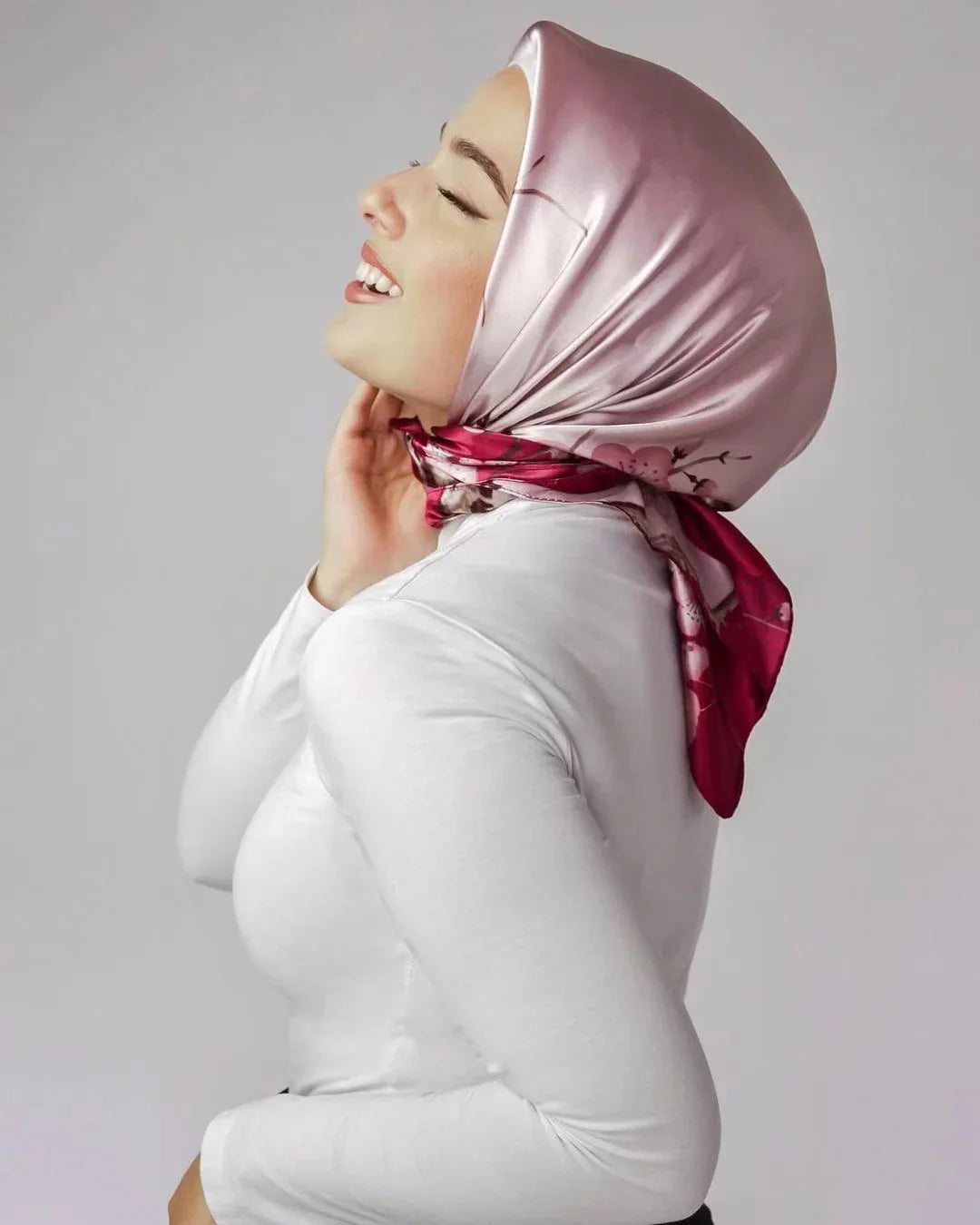 Smiling woman wearing a shiny pink floral silk hijab and white top against a plain background