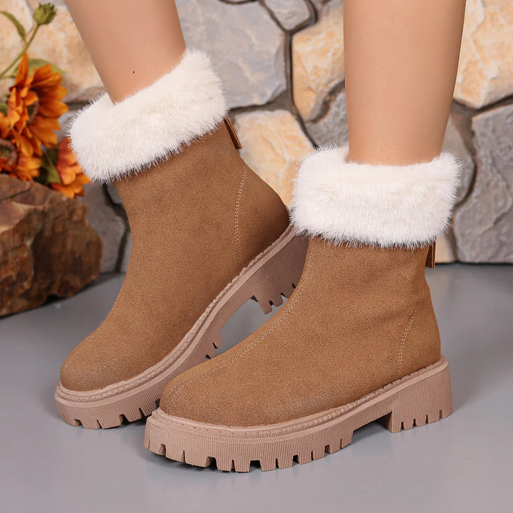 Brown suede ankle boots with white fur cuffs and chunky tread soles on a grey floor next to stone wall and orange flowers