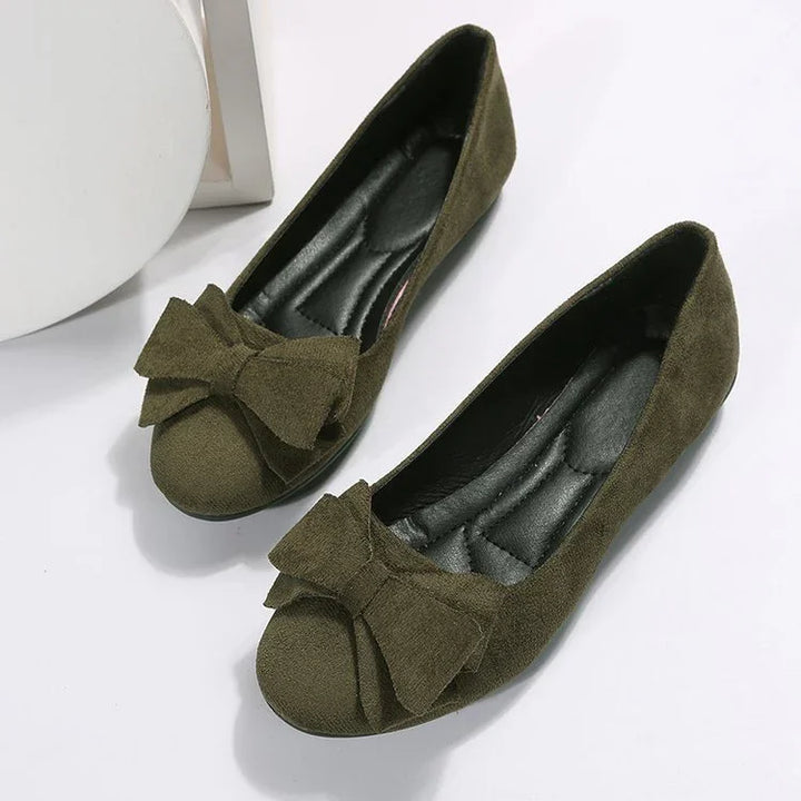Pair of olive green suede flat shoes with large bows on the toes on white background