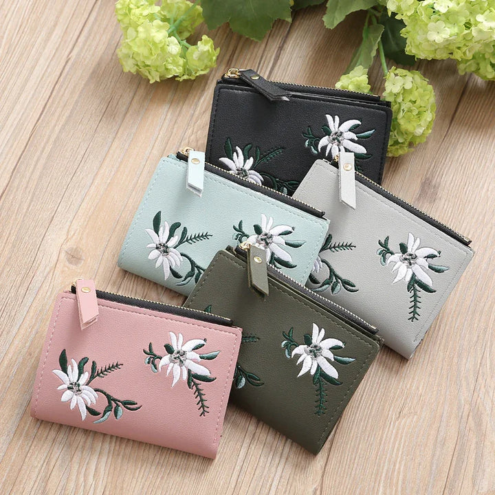Set of five small leather wallets in black, light blue, gray, pink, and olive with white floral embroidery, on wooden surface