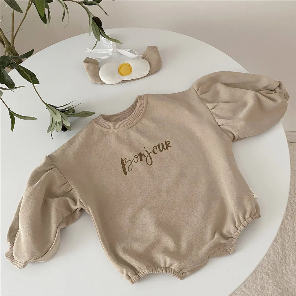 Beige baby romper with puff sleeves and 'Bonjour' print, displayed on a white round table with leafy plant and egg-shaped accessory