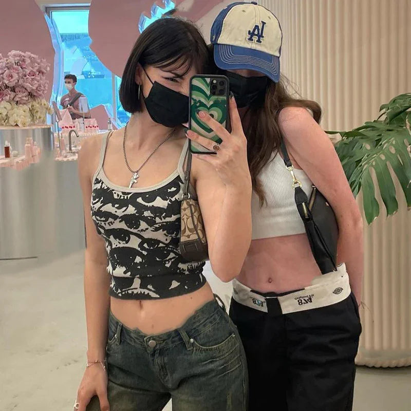 Two young women wearing black face masks and crop tops taking a mirror selfie indoors with plants and decor