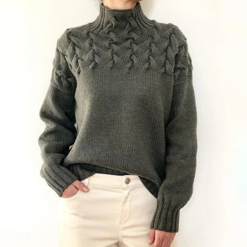 Person wearing green knitted turtleneck sweater with cable knit design and white pants
