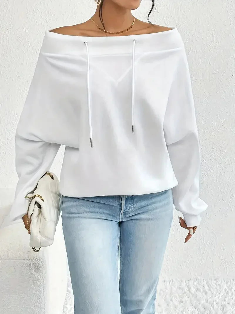 Woman wearing white off-shoulder long sleeve hoodie with drawstrings and light blue jeans holding white quilted bag