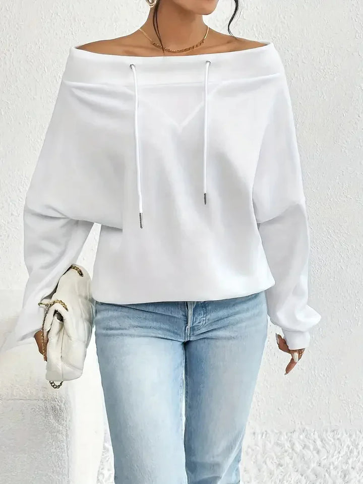 Woman wearing white off-shoulder long sleeve hoodie with drawstrings and light blue jeans holding white quilted bag