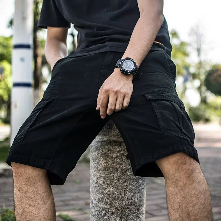 Man wearing black cargo shorts and a black watch sitting outdoors on a stone post