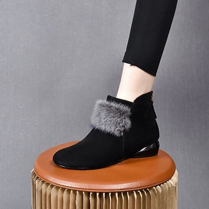 Black suede ankle boot with gray fur trim worn with black pants, displayed on a round brown stool