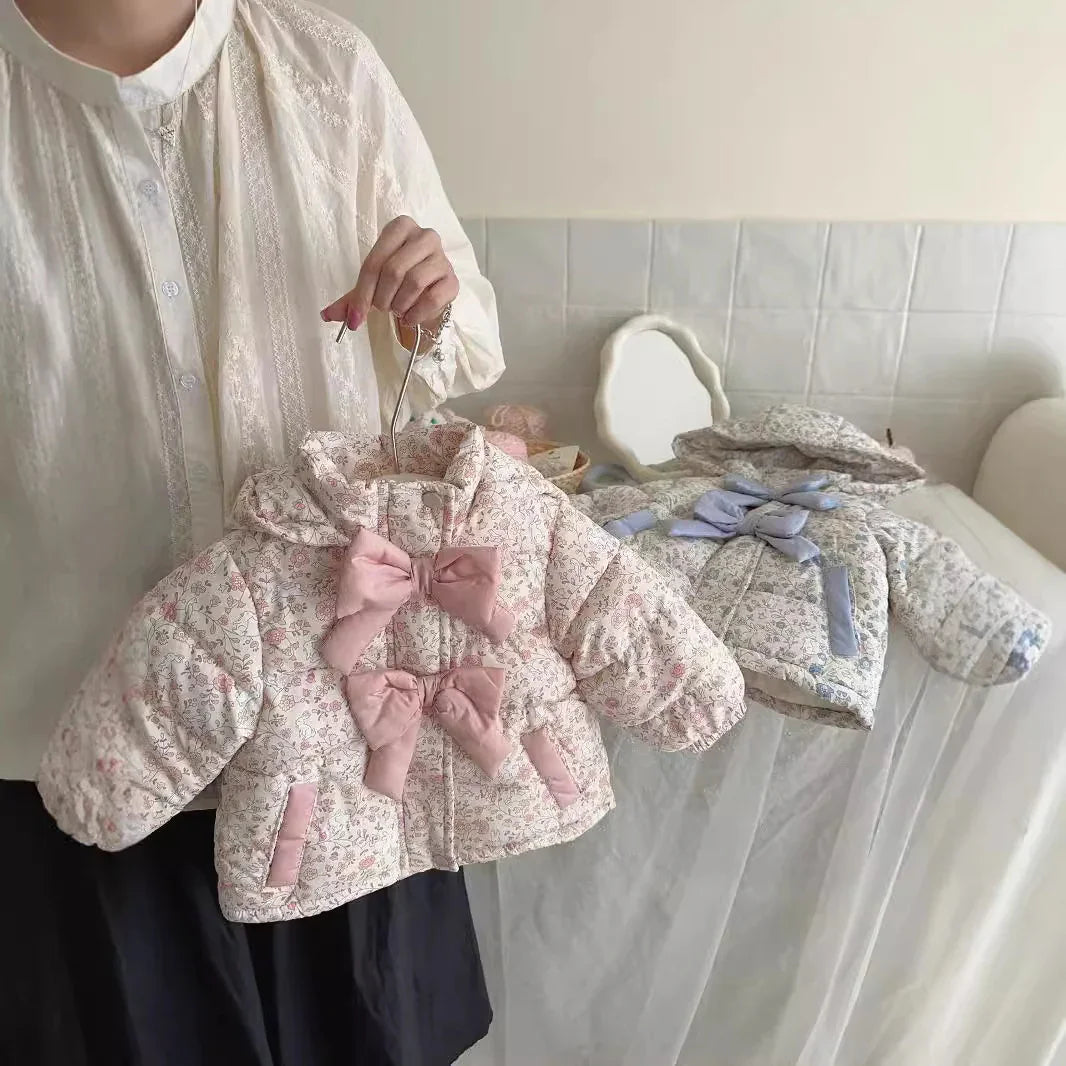 Person holding two floral patterned baby jackets with bows, in a light, cozy room setting