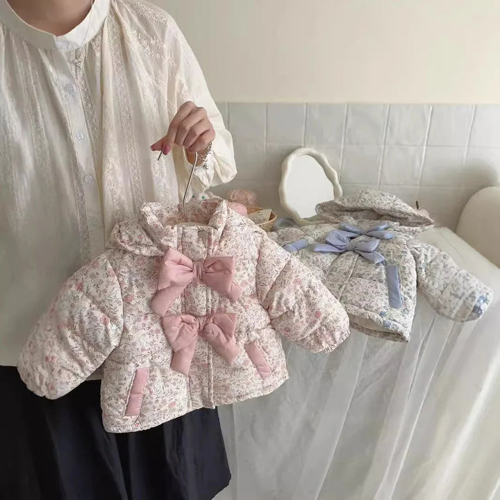 Person holding two floral patterned baby jackets with bows, in a light, cozy room setting