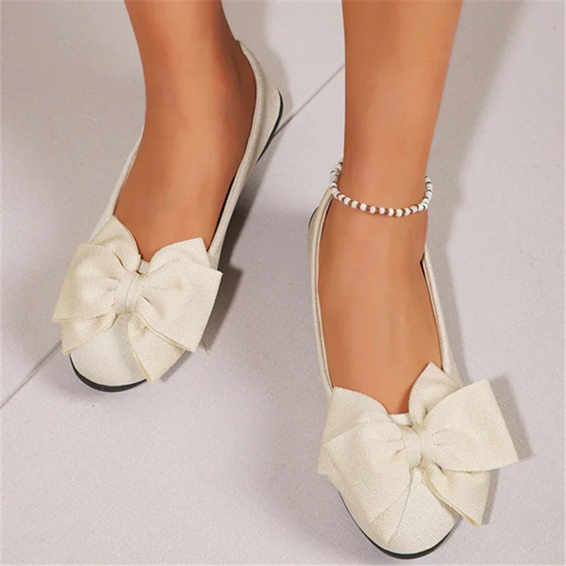 Cream textured ballet flats with large bows worn on feet on light tile floor
