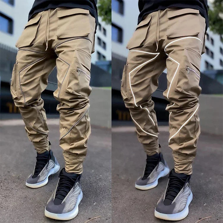 Men's beige cargo pants with zipper and reflective piping details worn with black and gray sneakers outdoors