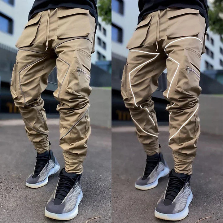 Men's beige cargo pants with zipper and reflective piping details worn with black and gray sneakers outdoors