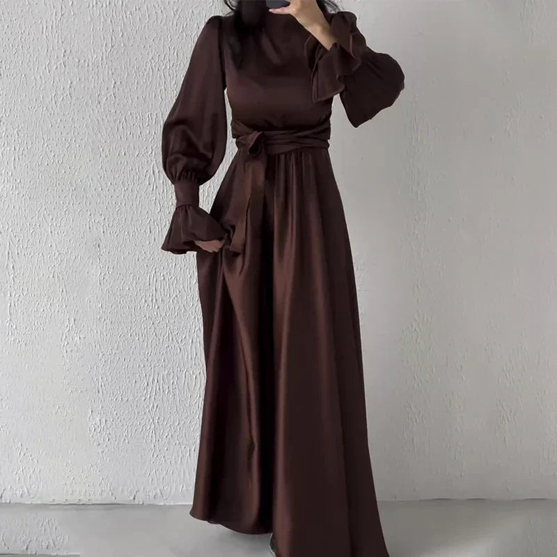 Woman wearing elegant dark brown long sleeve maxi dress with puffed sleeves and waist tie standing against white textured wall