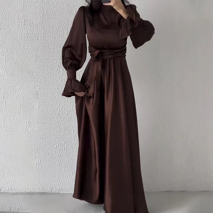 Woman wearing elegant dark brown long sleeve maxi dress with puffed sleeves and waist tie standing against white textured wall