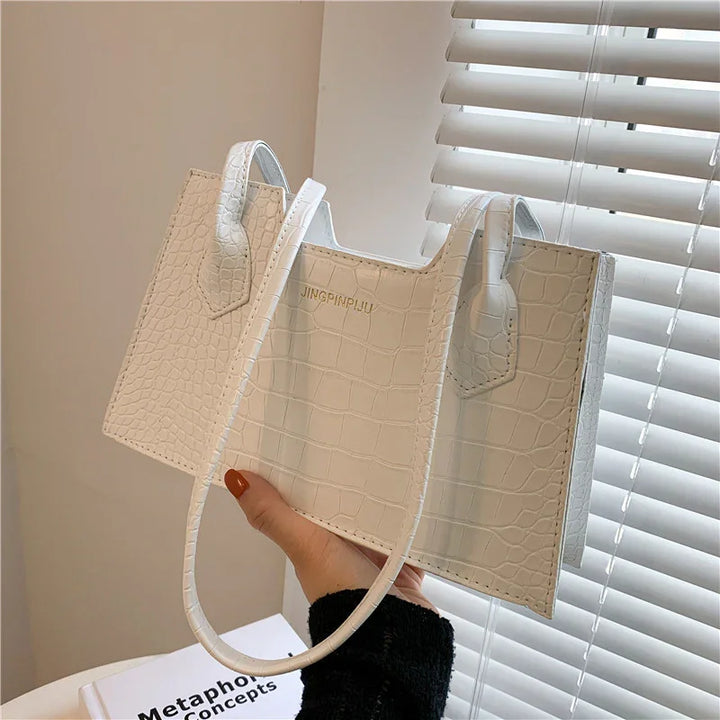 White crocodile texture handbag with handles held near window blinds over table with book