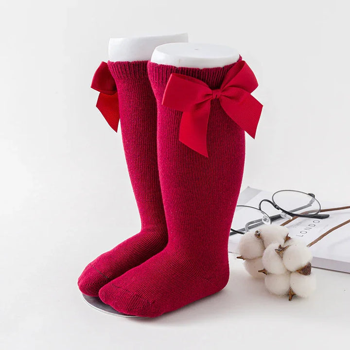 Pair of red knee-high baby socks with decorative bows on white display stands