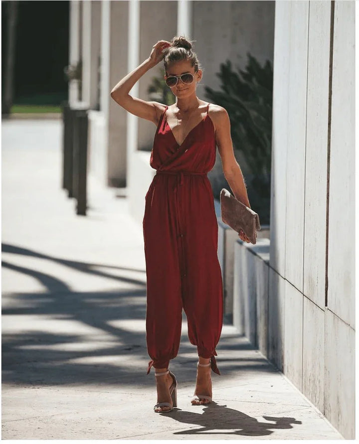Woman in red sleeveless jumpsuit with ankle ties and sunglasses walking outdoors in high heels holding a clutch