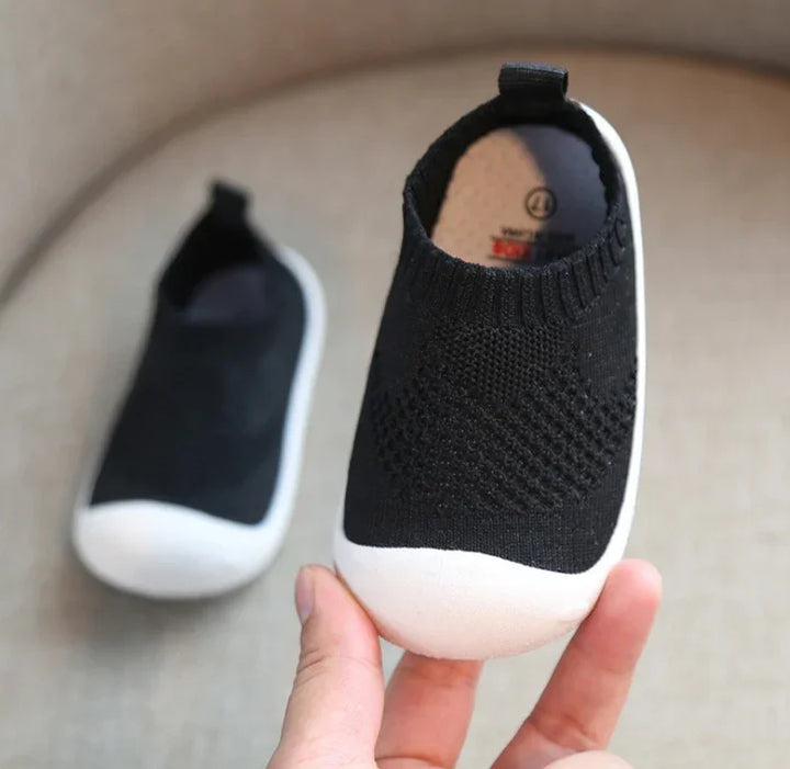 Black knit toddler slip-on shoes with white rubber soles held against beige background
