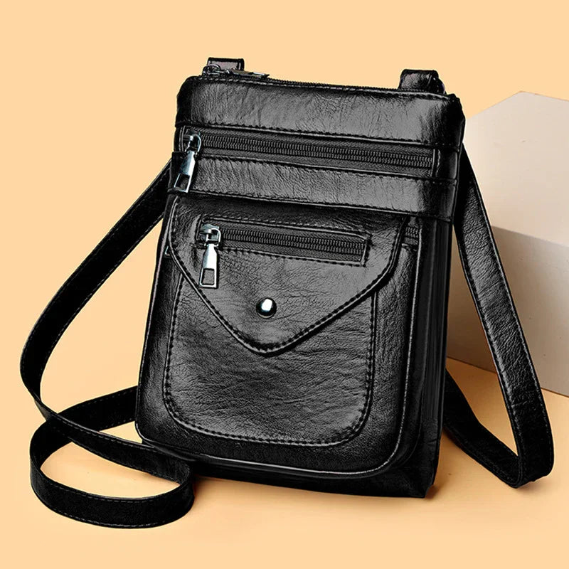 Black leather crossbody bag with zippered pockets and adjustable shoulder strap on beige background
