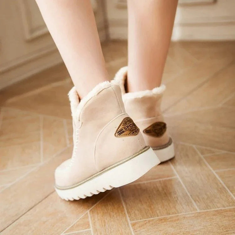 Beige winter boots with white fur lining worn on wooden herringbone floor, featuring brown logo patch