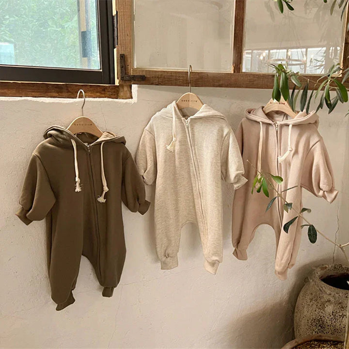 Three baby hooded zipper jumpsuits in brown, beige, and light pink hanging on wooden hangers near window