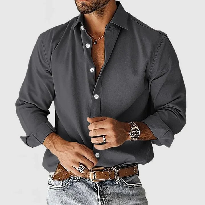 Man wearing dark gray button-up shirt with sleeves rolled, brown belt, light jeans, watch, rings, and necklace