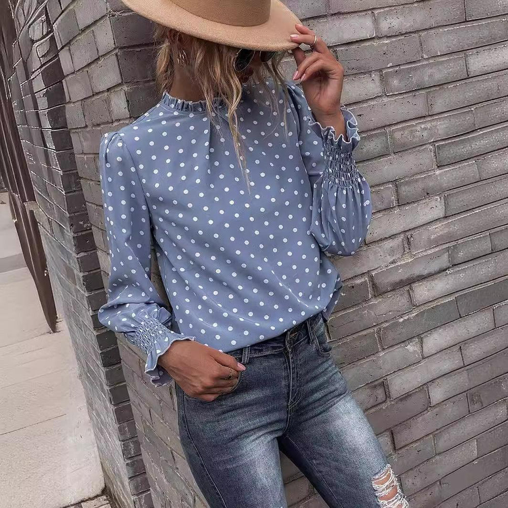 Woman wearing a blue polka dot blouse with ruffled cuffs and ripped jeans leaning against a gray brick wall
