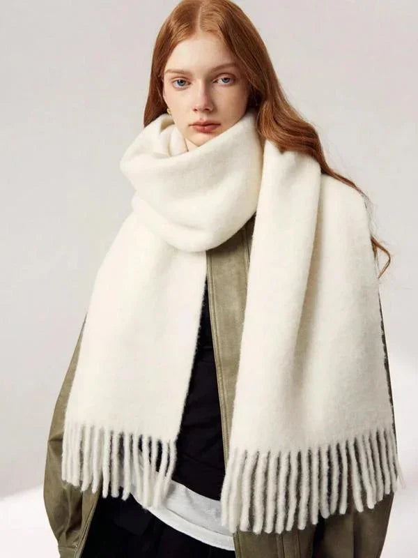 Young woman wearing a large white fringed wool scarf and olive green coat against a plain background