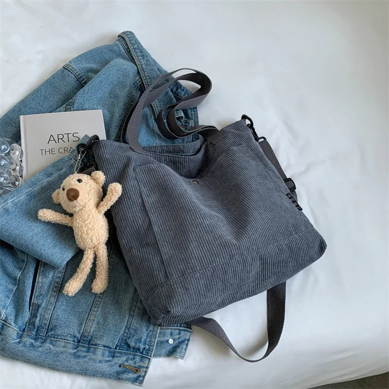 Gray corduroy tote bag with plush teddy bear keychain on denim jacket beside arts and crafts book