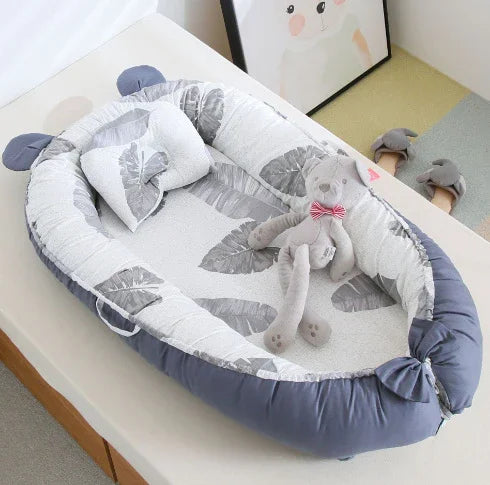 Baby nest bed with grey leaves pattern, matching pillow, and grey teddy bear with bow tie on a white surface