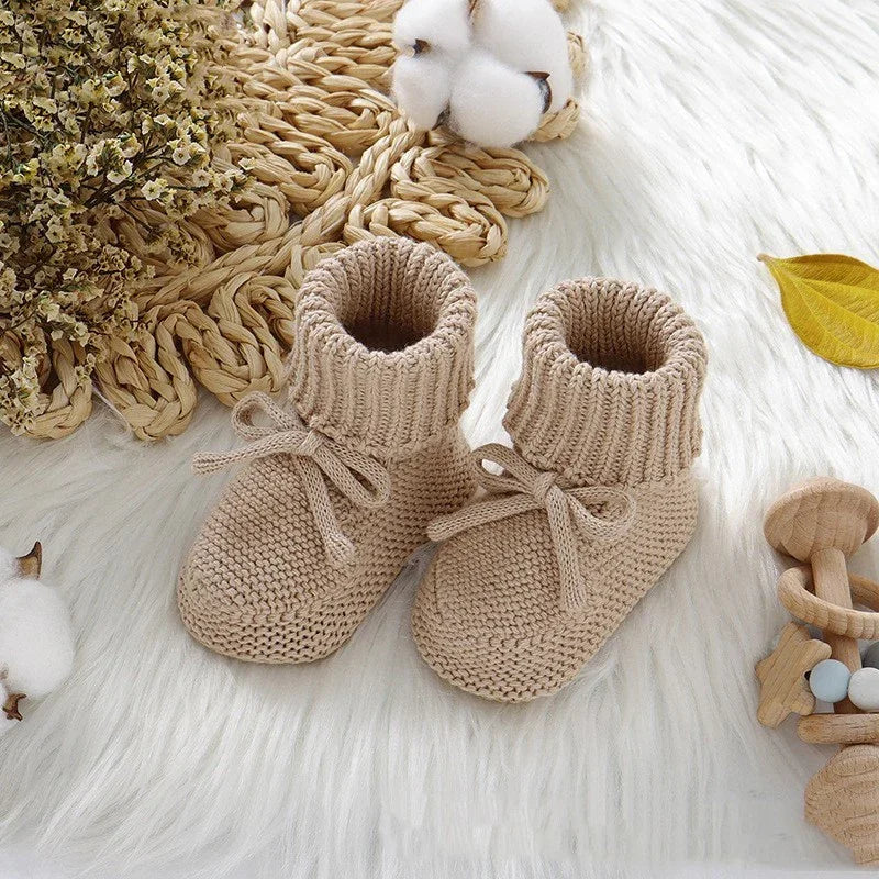 Beige knitted baby booties with laces on a white furry surface surrounded by cotton flowers and wooden toys