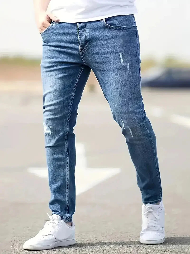 Man wearing slim fit blue distressed jeans and white sneakers standing outdoors on pavement.