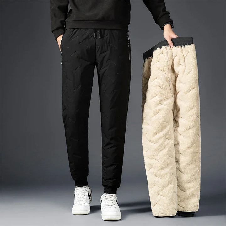 Man wearing black quilted jogger pants holding fleece-lined pants against gray background