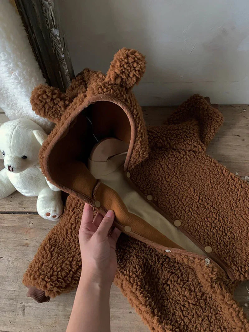 Hand holding open brown teddy bear hooded baby onesie on wooden floor with white teddy bear toy