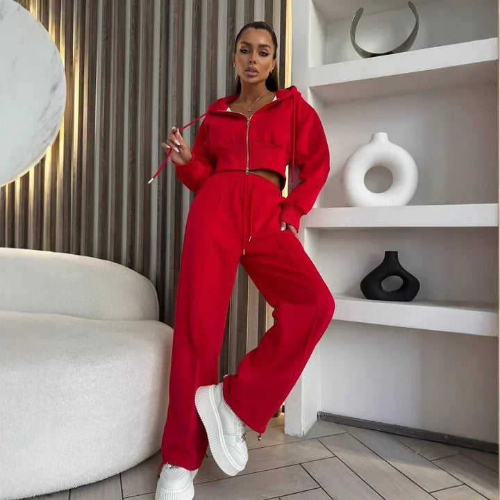 Woman in red cropped zip-up hoodie and matching wide-leg pants posing indoors by white couch and decorative shelves