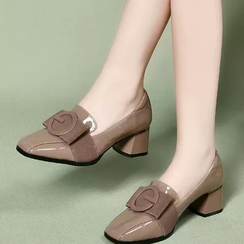 Women wearing taupe patent leather block heel shoes with a large bow and circular detail on top