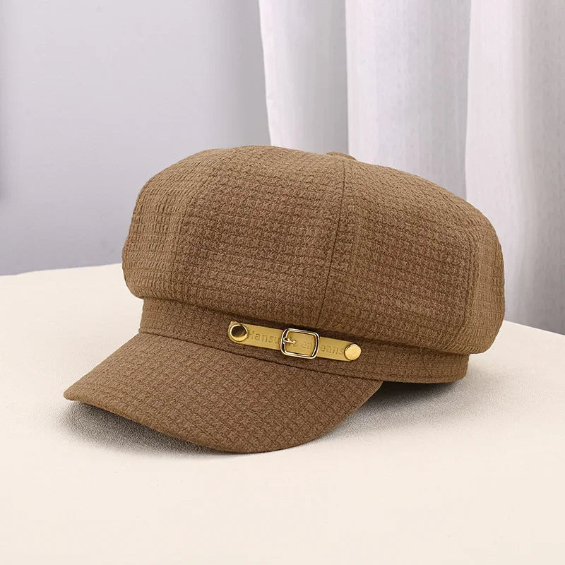 Brown textured newsboy cap with gold buckle detail on front visor, displayed on cream surface